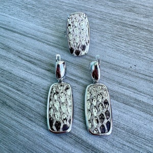 Może przedstawiać: Srebrny zestaw biżuterii składający się z pierścionka i pary kolczyków. Każdy element ma prostokątny kształt z teksturowanym wzorem przypominającym łuski w odcieniach bieli, szarości i czerni. Kolczyki są wiszące.