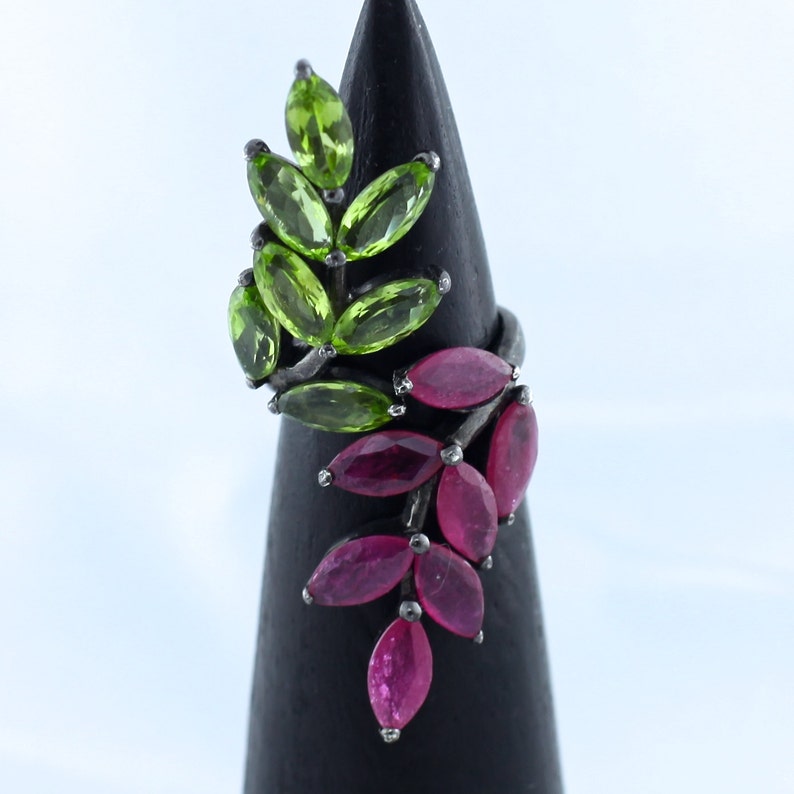 Pu&ograve; includere: Un anello d'argento con un design a foglie con pietre preziose verdi e rosa. L'anello ha una fascia in argento scuro e le pietre sono disposte per assomigliare a foglie. Le pietre sono ovali e variano dal verde chiaro al rosa.