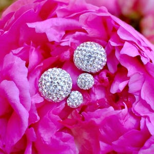 May include: Four spherical stud earrings of varying sizes, each covered in clear, reflective crystals that catch the light, resting against the vibrant, layered petals of a bright pink peony flower in full bloom.