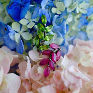 May include: A close-up of two gemstone rings with a floral design. One ring features green gemstones, the other features pink gemstones. The rings are set against a backdrop of blue, pink, and white flowers.
