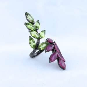 Pu&ograve; includere: Anello in argento con design a foglie ornato da pietre preziose verdi e rosse. L'anello ha una finitura argento scuro e le pietre preziose ovali sono disposte per imitare le foglie. Sfondo bianco.