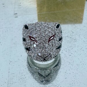 Puede incluir: Un anillo de plata con un dise&ntilde;o de cara de tigre en blanco y negro. Los ojos del tigre son rojos y el anillo est&aacute; cubierto de peque&ntilde;os diamantes de imitaci&oacute;n transparentes.