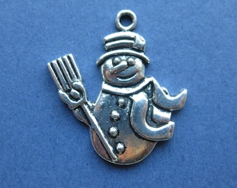 May include: A silver-colored snowman charm with a broom, hat, and scarf. The snowman has a smiling face and is made of metal. The charm has a loop at the top for attaching to a necklace or bracelet. The background is a solid blue color.
