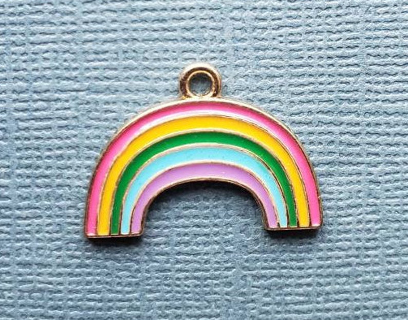 K&ouml;nnte beinhalten: Ein goldfarbener Regenbogen-Anh&auml;nger mit pinkfarbener, gelber, gr&uuml;ner, blauer und violetter Emaille-Oberfl&auml;che. Der Anh&auml;nger ist in Form eines Regenbogenbogens.