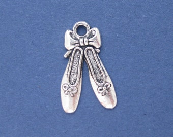 Könnte beinhalten: Silberfarbener Metallanhänger mit einem Paar Ballettschuhen mit Schleifen. Der Anhänger hat oben eine Schlaufe zum Befestigen an einer Halskette oder einem Armband. Die Ballettschuhe sind mit strukturierten Akzenten versehen.
