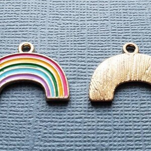 K&ouml;nnte beinhalten: Zwei goldfarbene Regenbogen-Anh&auml;nger. Ein Anh&auml;nger ist ein Regenbogen mit roter, orangefarbener, gelber, gr&uuml;ner, blauer, indigofarbenem und violetter Emaille. Der andere Anh&auml;nger ist ein einfarbiger goldfarbener Regenbogen.
