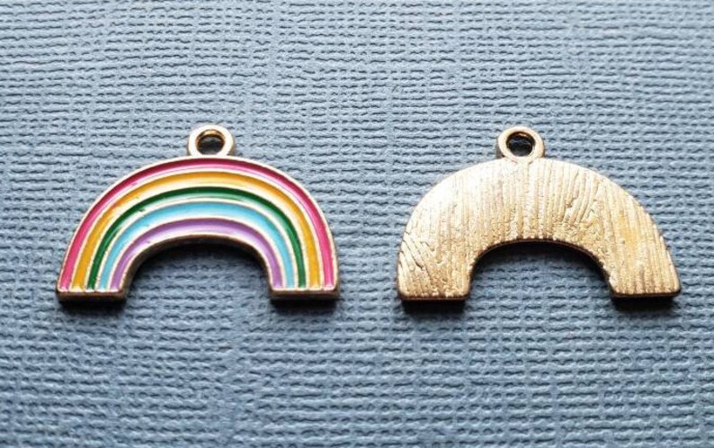 K&ouml;nnte beinhalten: Zwei goldfarbene Regenbogen-Anh&auml;nger. Ein Anh&auml;nger ist ein Regenbogen mit roter, orangefarbener, gelber, gr&uuml;ner, blauer, indigofarbenem und violetter Emaille. Der andere Anh&auml;nger ist ein einfarbiger goldfarbener Regenbogen.