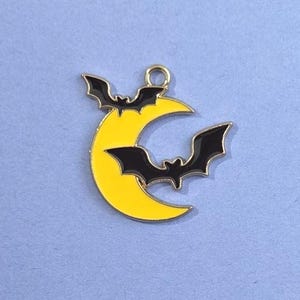 May include: A gold-colored metal charm featuring a yellow crescent moon with two black bats. The charm has a small loop at the top for attaching to a necklace or bracelet. The design is suitable for Halloween-themed jewelry.