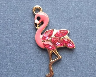 May include: A pink flamingo charm with gold-colored accents. The flamingo has a black eye and is adorned with pink crystal-like embellishments on its wings. The charm has a loop at the top for attaching to a necklace or bracelet.