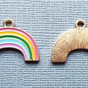 K&ouml;nnte beinhalten: Zwei goldfarbene Regenbogen-Anh&auml;nger. Ein Anh&auml;nger ist ein Regenbogen mit rosa, gelber, gr&uuml;ner, blauer und wei&szlig;er Emaille. Der andere Anh&auml;nger ist ein durchg&auml;ngig goldfarbener Regenbogen.