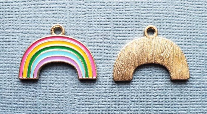 K&ouml;nnte beinhalten: Zwei goldfarbene Regenbogen-Anh&auml;nger. Ein Anh&auml;nger ist ein Regenbogen mit rosa, gelber, gr&uuml;ner, blauer und wei&szlig;er Emaille. Der andere Anh&auml;nger ist ein durchg&auml;ngig goldfarbener Regenbogen.
