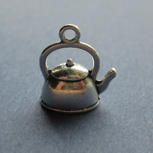 May include: A small, silver-colored kettle charm with a loop at the top for hanging. The charm has a handle, spout, and a small knob on the lid. The kettle is a miniature version of a classic design.