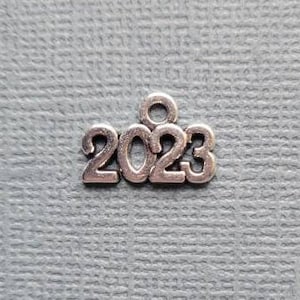 May include: Silver-colored metal charm with the numbers "2023" in a sans-serif font. The charm has a small loop at the top for attaching to a necklace or bracelet. The background is a textured, light blue surface.