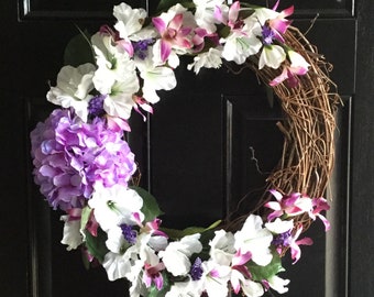 May include: A grapevine wreath decorated with white, purple and pink artificial flowers. The wreath is hanging on a black door.
