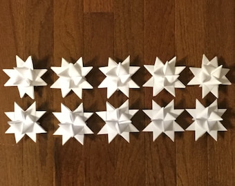 May include: Ten white paper star ornaments arranged in two rows on a wooden surface. Each star has multiple points, creating a symmetrical design. The stars are evenly spaced, and the wood grain provides a warm background.