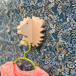 May include: A wooden animal head shaped like a lion with a green hanger attached to it. The hanger is hanging on a wall with a floral pattern wallpaper.