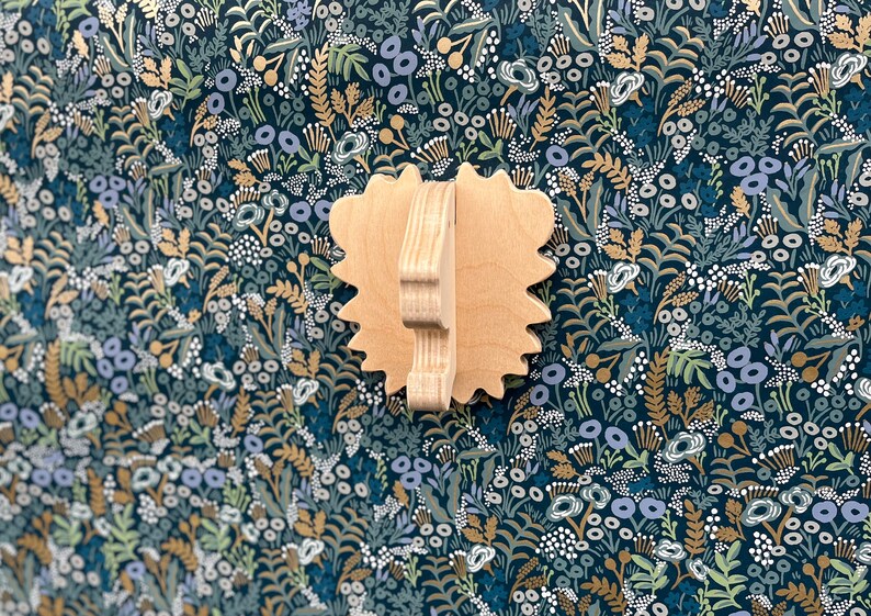 May include: A wooden wall-mounted decorative piece shaped like a lion's head with a textured surface. The piece is painted in a light brown color and is set against a dark blue background with a floral pattern.