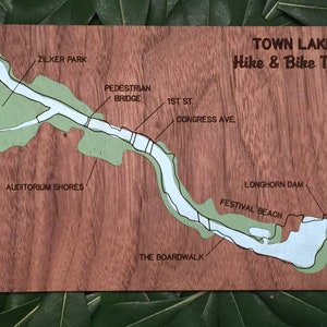 May include: A wooden map of Town Lake Hike & Bike Trail in Austin, Texas. The map shows the trail winding around the lake, with labels for Zilker Park, Auditorium Shores, Pedestrian Bridge, 1st St, Congress Ave, The Boardwalk, Longhorn Dam, and Festival Beach.