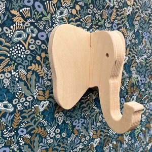 Puede incluir: Un gancho de pared de madera con forma de elefante con un acabado marr&oacute;n claro. El gancho tiene forma de cabeza de elefante con una trompa que se curva hacia abajo para formar un gancho.