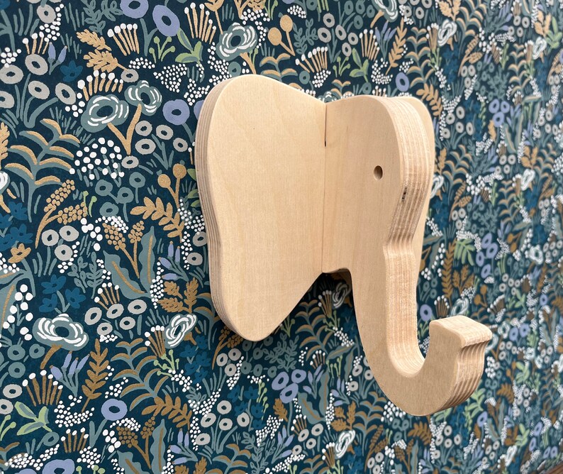 Puede incluir: Un gancho de pared de madera con forma de elefante con un acabado marr&oacute;n claro. El gancho tiene forma de cabeza de elefante con una trompa que se curva hacia abajo para formar un gancho.