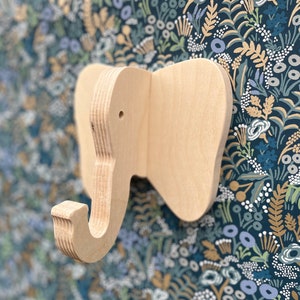 Puede incluir: Un gancho de pared de madera con forma de elefante con un acabado natural. El gancho est&aacute; montado en un papel tapiz floral azul y dorado.