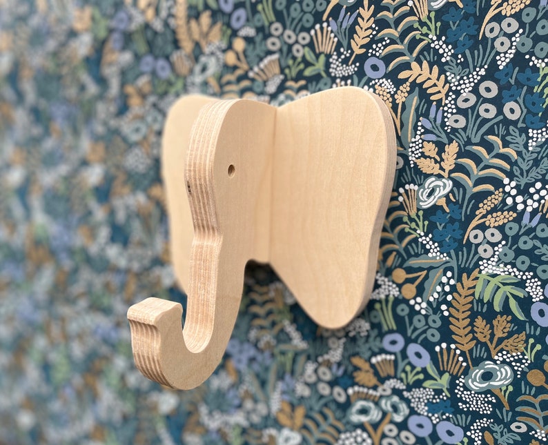 Puede incluir: Un gancho de pared de madera con forma de elefante con un acabado natural. El gancho est&aacute; montado en un papel tapiz floral azul y dorado.