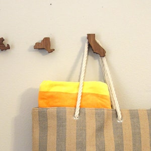 May include: Three wooden state-shaped hooks mounted on a wall. The hooks are shaped like Massachusetts, New York, and California. A striped tote bag with a yellow and orange towel inside is hanging from the California hook. A blue plastic shovel is leaning against the wall.