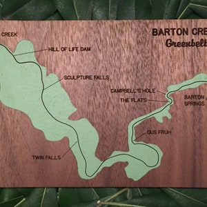 May include: A wooden map of Barton Creek Greenbelt, a trail in Austin, Texas. The map shows the trail's path, along with the names of various points of interest, including Lost Creek, Hill of Life Dam, Twin Falls, Sculpture Falls, Campbell's Hole, The Flats, Barton Springs, and Gus Fruh.