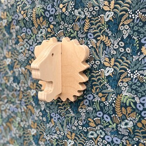 May include: A wooden lion head wall decor with a light brown finish. The lion head is carved with detailed features and has a textured surface. The background is a dark blue floral pattern.