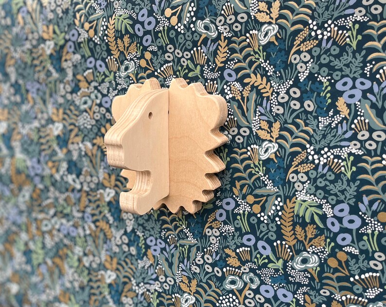 May include: A wooden lion head wall decor with a light brown finish. The lion head is carved with detailed features and has a textured surface. The background is a dark blue floral pattern.