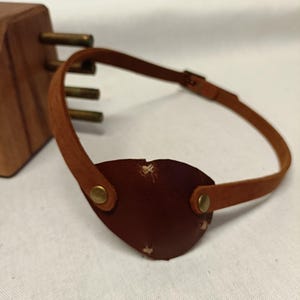 Puede incluir: Un parche para el ojo de cuero marrón con una correa y remaches de latón. El parche para el ojo tiene forma ovalada y tiene un pequeño desgarro. La correa es ajustable y se conecta al parche para el ojo con remaches de latón.