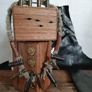 Może przedstawiać: Drewniana rzeźba z naszyjnikiem z sznurka wykonanym z łusek od strzelby. Rzeźba składa się z dwóch drewnianych bloków, jeden na drugim. Górny blok ma cztery łuski od strzelby wystające z niego. Dolny blok ma trzy łuski od strzelby przymocowane do naszyjnika z sznurka. Rzeźba stoi na drewnianej powierzchni.