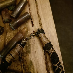 May include: A handcrafted necklace featuring a black cord, adorned with tan twine and a bronze clasp. The necklace is presented on a wooden surface, accompanied by several brass bullet casings, offering a rustic, industrial style.