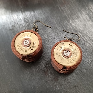 May include: A pair of handmade earrings featuring repurposed 12 GA Winchester shotgun shell casings. The earrings have a brown leather backing and brass-colored metal accents. The text "WINCHESTER" and "12 GA" are visible on the casings.