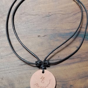 May include: A round, light brown pendant necklace with a black cord. The pendant has etched symbols and the word "Amillennium". The cord is knotted at the top and hangs on a wooden surface.