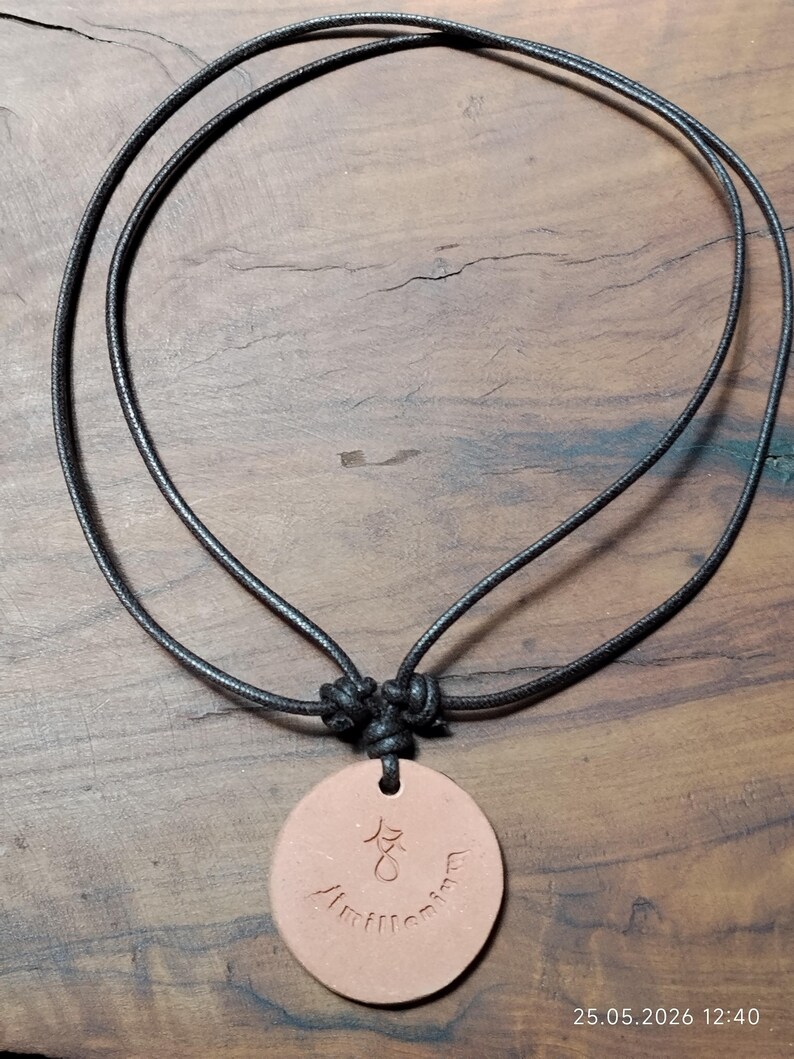 May include: A round, light brown pendant necklace with a black cord. The pendant has etched symbols and the word "Amillennium". The cord is knotted at the top and hangs on a wooden surface.