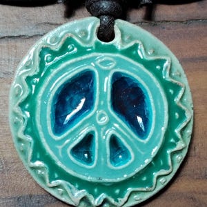 May include: A ceramic peace sign pendant in shades of teal and blue, strung on a black leather cord. The pendant has a textured, glazed finish and a decorative border. The peace symbol is centered within the pendant.