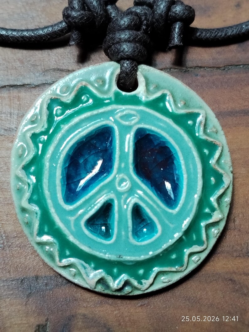 May include: A ceramic peace sign pendant in shades of teal and blue, strung on a black leather cord. The pendant has a textured, glazed finish and a decorative border. The peace symbol is centered within the pendant.