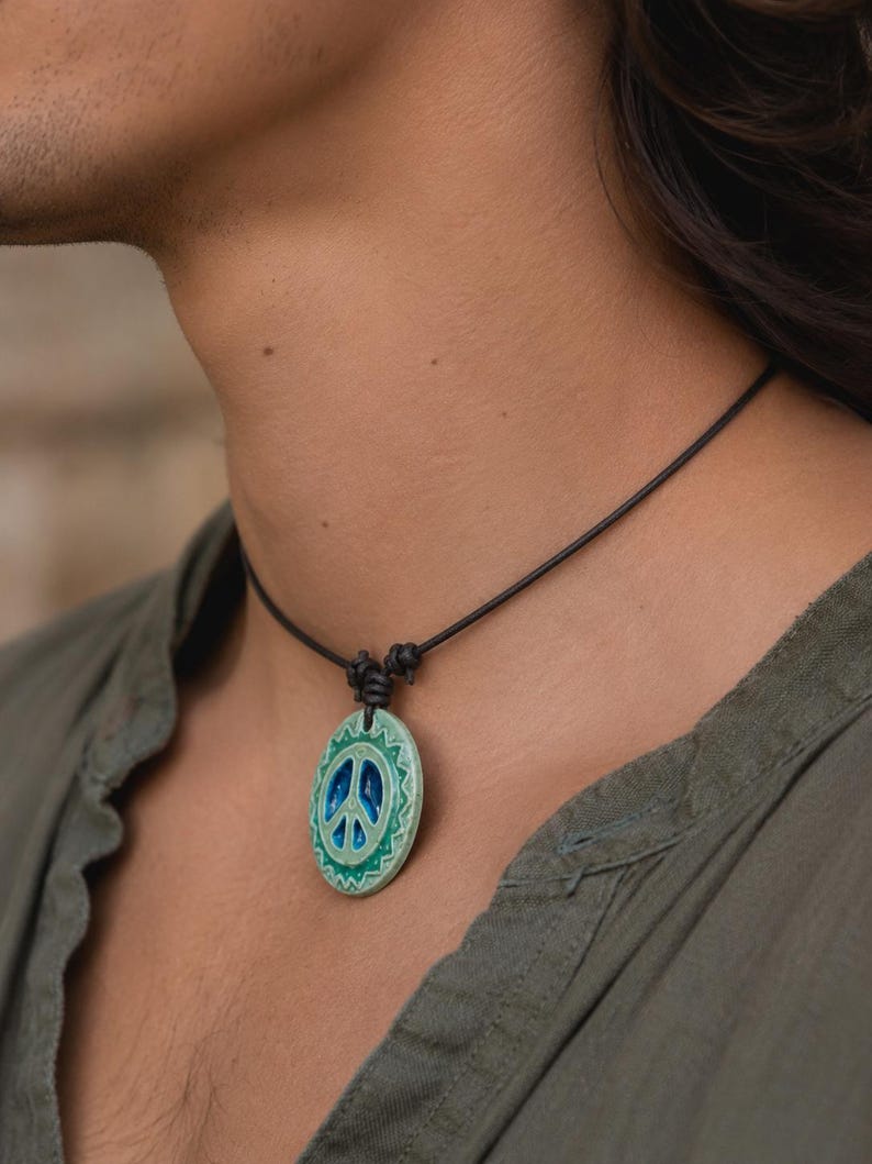 May include: A peace sign pendant necklace. The pendant is oval-shaped with a blue peace symbol in the center, surrounded by a green and white design. The necklace has a black cord.
