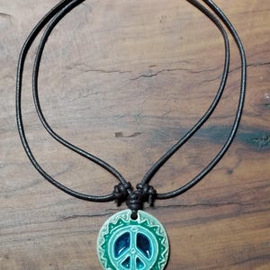 May include: A ceramic peace sign pendant necklace with a dark cord. The pendant features a teal and blue peace symbol with a decorative border. The necklace is displayed on a wooden surface.