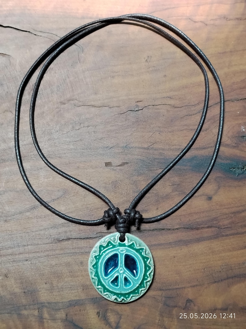 May include: A ceramic peace sign pendant necklace with a dark cord. The pendant features a teal and blue peace symbol with a decorative border. The necklace is displayed on a wooden surface.