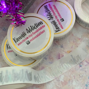May include: Three rolls of white washi tape with gold trim. The tape has a silver foil design that says "Kawaii Addiction" and a website address. The tape is on a white surface with a purple and silver garland in the background.