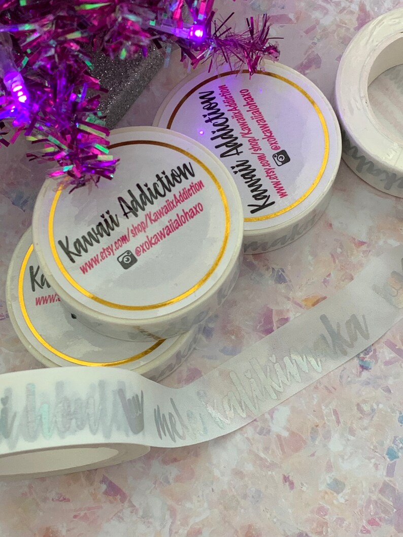 May include: Three rolls of white washi tape with gold trim. The tape has a silver foil design that says "Kawaii Addiction" and a website address. The tape is on a white surface with a purple and silver garland in the background.