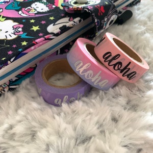 May include: Three rolls of washi tape with a gradient color design. The tape is pink, purple, and blue. The word "Aloha" is written in silver on each roll.