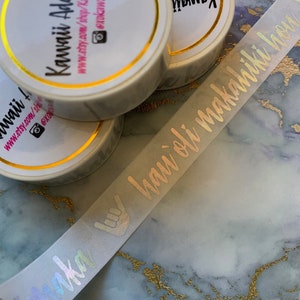 May include: A close-up of a white washi tape roll with gold accents. The tape has a holographic finish and features the text "Aloha" in a script font. The tape is on a marble surface.