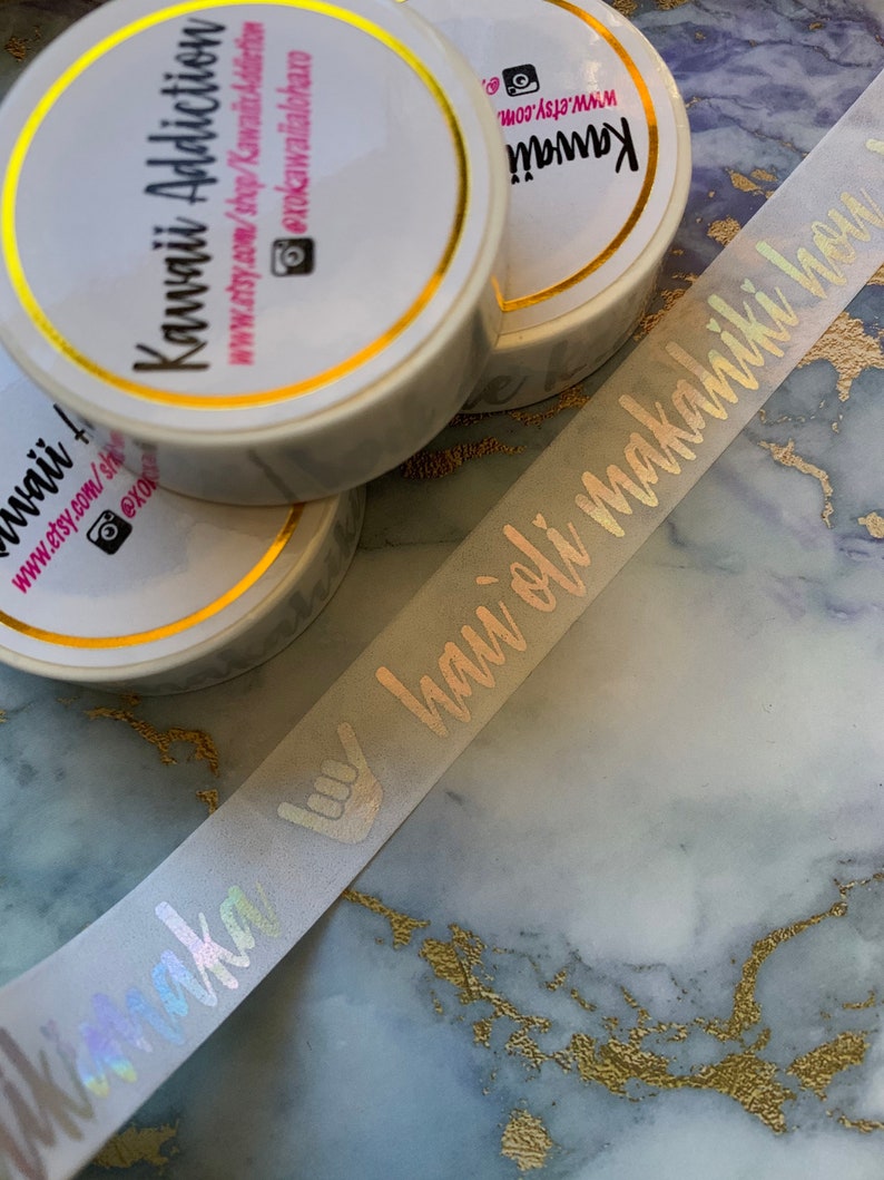 May include: A close-up of a white washi tape roll with gold accents. The tape has a holographic finish and features the text "Aloha" in a script font. The tape is on a marble surface.