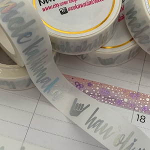 May include: A roll of white washi tape with a silver holographic design that says "hau oli maikahiki hou". The tape is wrapped around a roll with a gold trim and the text "Kawaii Addiction" with a website and social media handle.