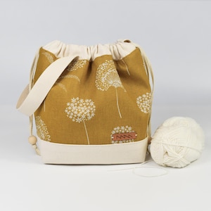 Op de afbeelding: Mosterdgele stoffen tas met een wit paardenbloemmotief. De tas heeft een crèmekleurige basis, een trekkoordsluiting en een bijpassende handgreep. Een bolletje witte wol ligt naast de tas.