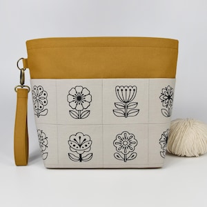 May include: Mustard yellow and cream fabric pouch featuring a black floral design. The pouch has a matching wrist strap and a metal clasp. A ball of yarn sits beside the pouch. The pouch is designed for storing craft supplies.