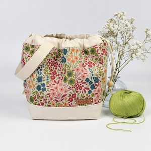 Puede incluir: Una bolsa de hilo con estampado floral con base, asa y cierre de cordón de color crema. La bolsa presenta un colorido estampado de flores en rojo, azul, naranja y verde. Una madeja de hilo verde está al lado de la bolsa.